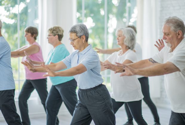 A multi-ethnic group of senior adults are taking a tai chi fitness class. They are practicing their martial art inside of a brightly lit studio. Here, a woman of asian ethnicity is leading the class through movements.